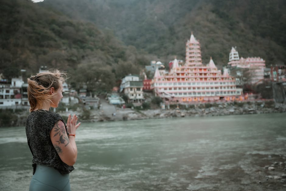 A woman practices yoga by the Ganges River in Rishikesh with a temple in the background.